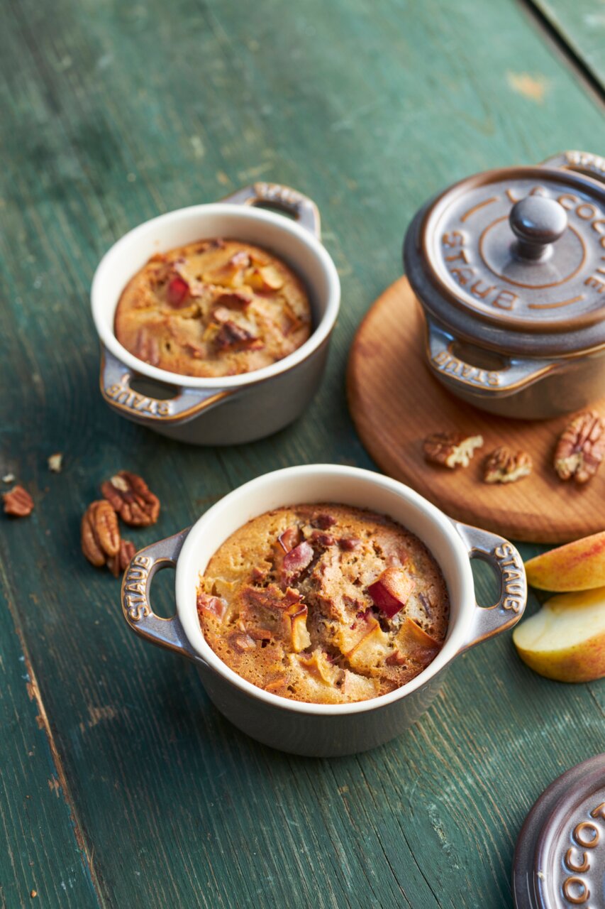 Zwei graue Mini-Cocottes mit gebackenem Dessert, umgeben von Pekannüssen und Apfelscheiben, aus der Vogelperspektive fotografiert.