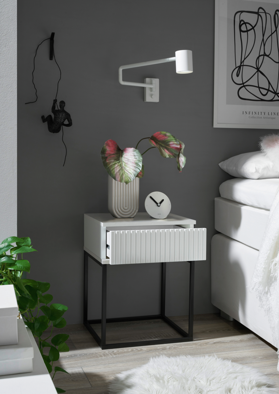 Moderne Nachtkommode mit weißer Schublade und schwarzem Metallgestell, dekoriert mit einer Pflanze und einer Uhr, seitliche Perspektive.
