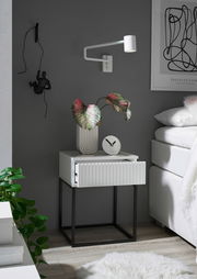 Moderne Nachtkommode mit weißer Schublade und schwarzem Metallgestell, dekoriert mit einer Pflanze und einer Uhr, seitliche Perspektive.