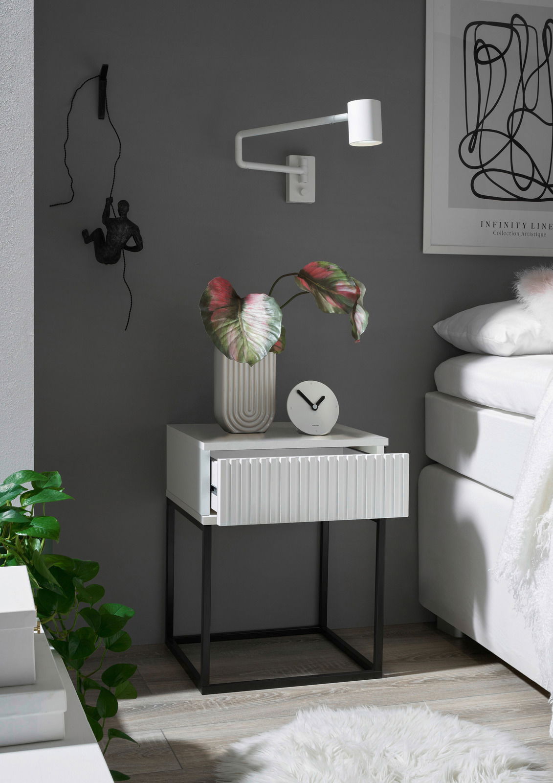 Moderne Nachtkommode mit weißer Schublade und schwarzem Metallgestell, dekoriert mit einer Pflanze und einer Uhr, seitliche Perspektive.