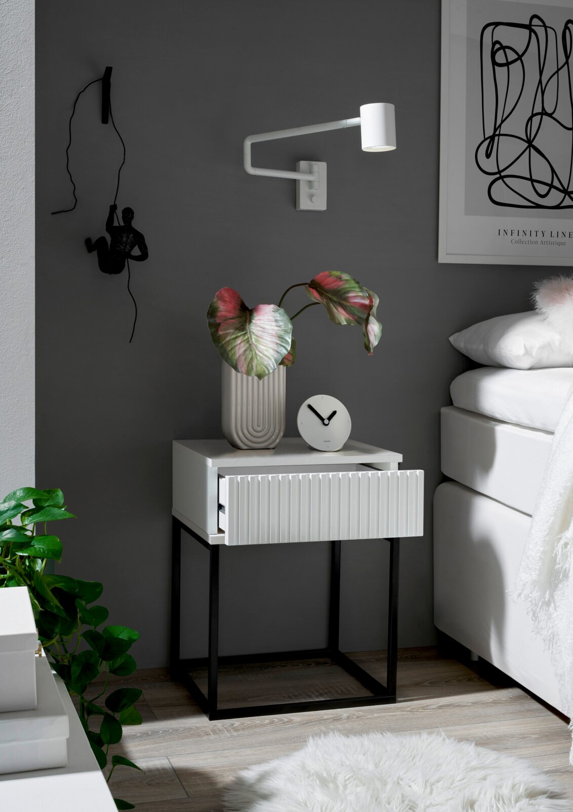 Moderne Nachtkommode mit weißer Schublade und schwarzem Metallgestell, dekoriert mit einer Pflanze und einer Uhr, seitliche Perspektive.
