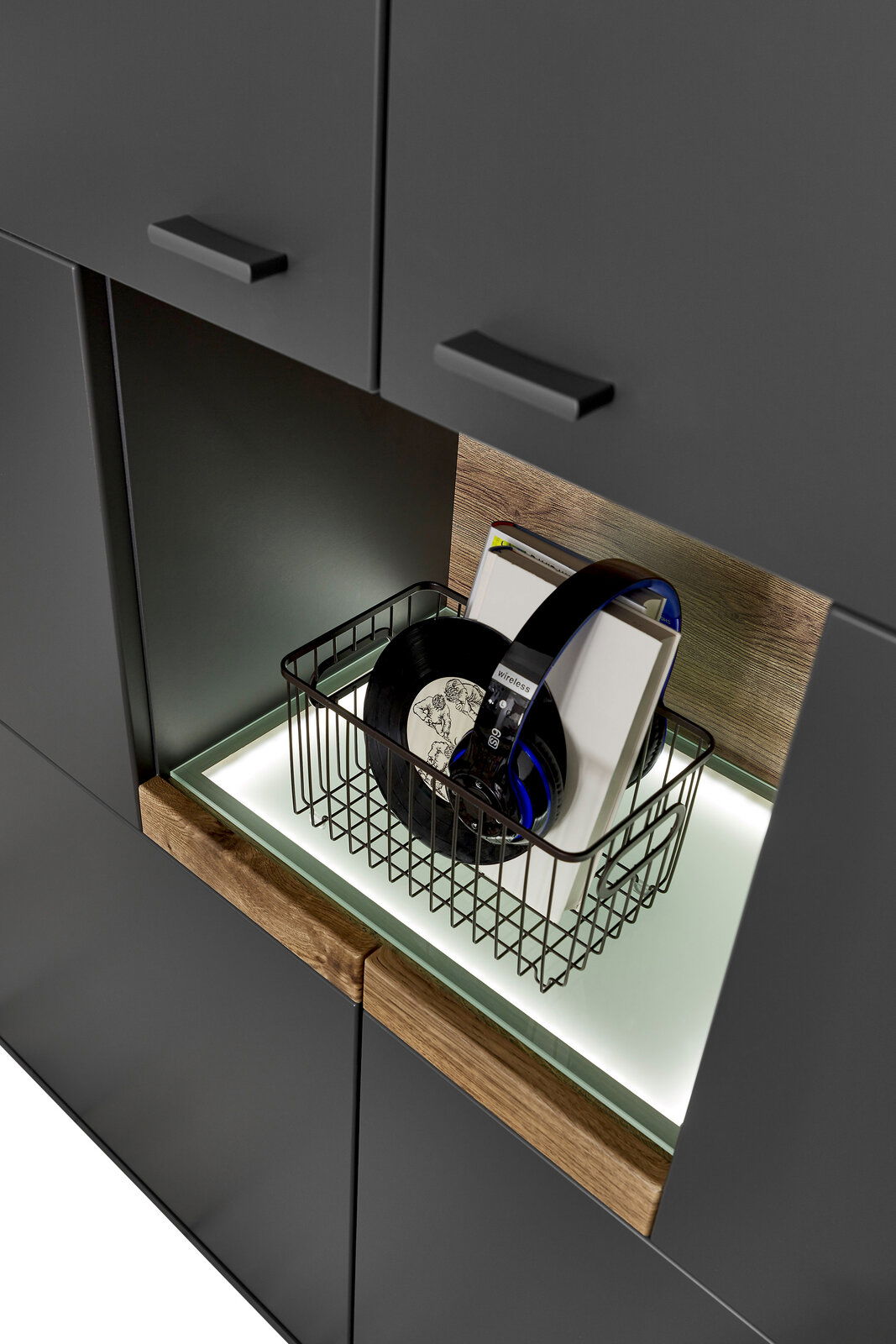 Nahaufnahme eines modernen Highboards mit dunklen Schranktüren und einem offenen Fach mit Beleuchtung, das einen Korb mit Schallplatten und Kopfhörern enthält. Perspektive von schräg oben.