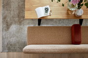 Draufsicht auf eine moderne Wohnzimmerecke mit Holz-Couchtisch, offenem Notizbuch, Glas und Blumen sowie beigefarbenem Sofa mit rostfarbenem Kissen auf grauem Teppich.