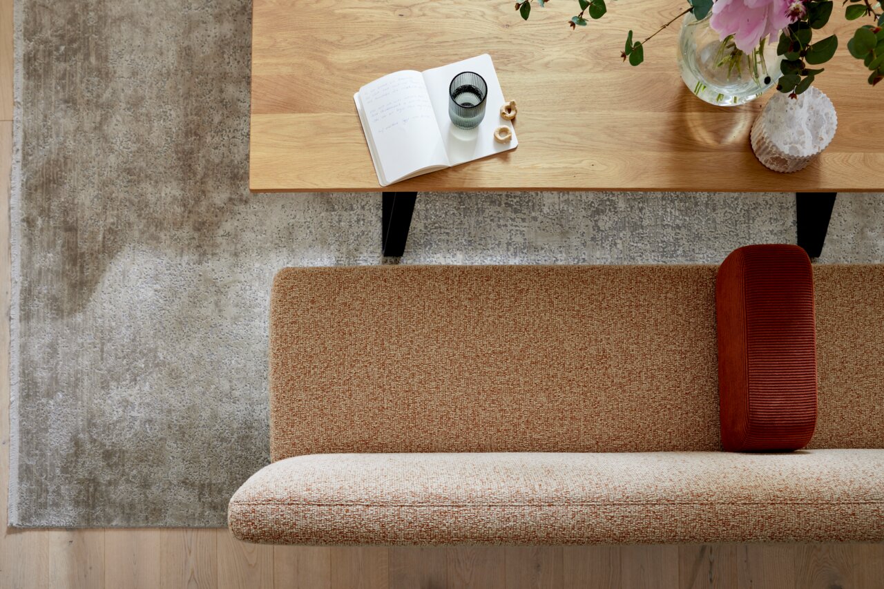 Bovenaanzicht van een moderne zithoek met een houten salontafel, een open notitieboekje, een glas en bloemen, evenals een beige bank met roestkleurige kussens op een grijs tapijt.