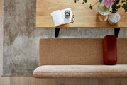 Bovenaanzicht van een moderne zithoek met een houten salontafel, een open notitieboekje, een glas en bloemen, evenals een beige bank met roestkleurige kussens op een grijs tapijt.