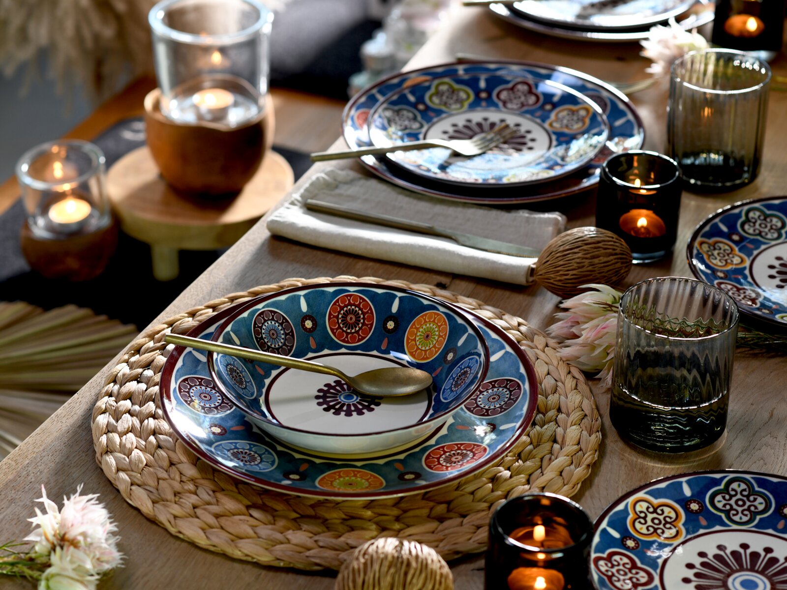 Schräg von oben fotografierte, stilvoll gedeckte Tafel mit bunt gemustertem Teller- und Schüssel-Set auf geflochtenem Platzset, goldfarbenem Besteck, Kerzen und geriffelten Gläsern auf Holztisch.