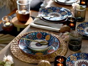 Schräg von oben fotografierte, stilvoll gedeckte Tafel mit bunt gemustertem Teller- und Schüssel-Set auf geflochtenem Platzset, goldfarbenem Besteck, Kerzen und geriffelten Gläsern auf Holztisch.