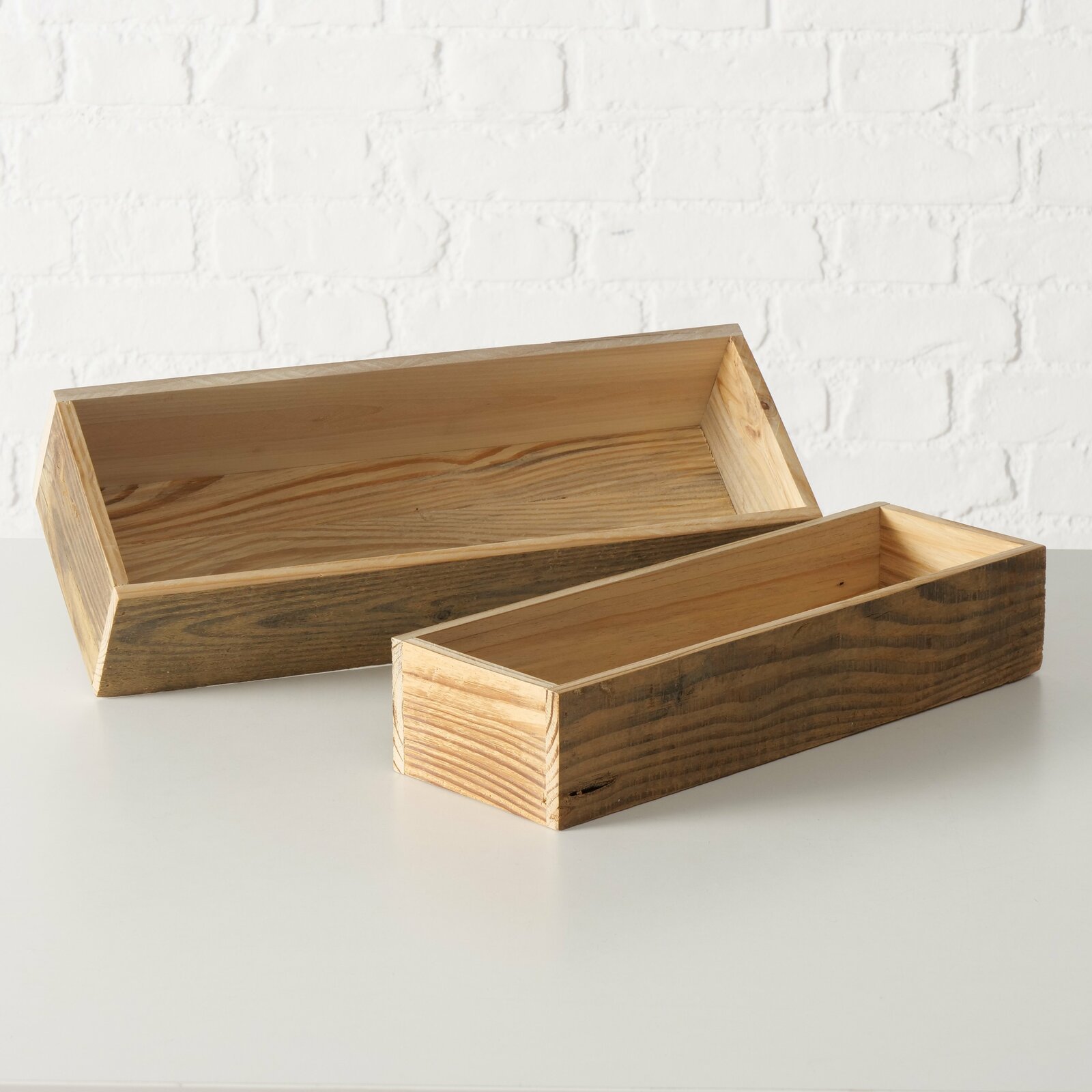 Zwei rechteckige Holzkisten, eine größer und eine kleiner, auf einer weißen Oberfläche vor einer weißen Ziegelwand. Die Kisten sind aus hellem Holz mit sichtbarer Maserung gefertigt. Perspektive von vorne.
