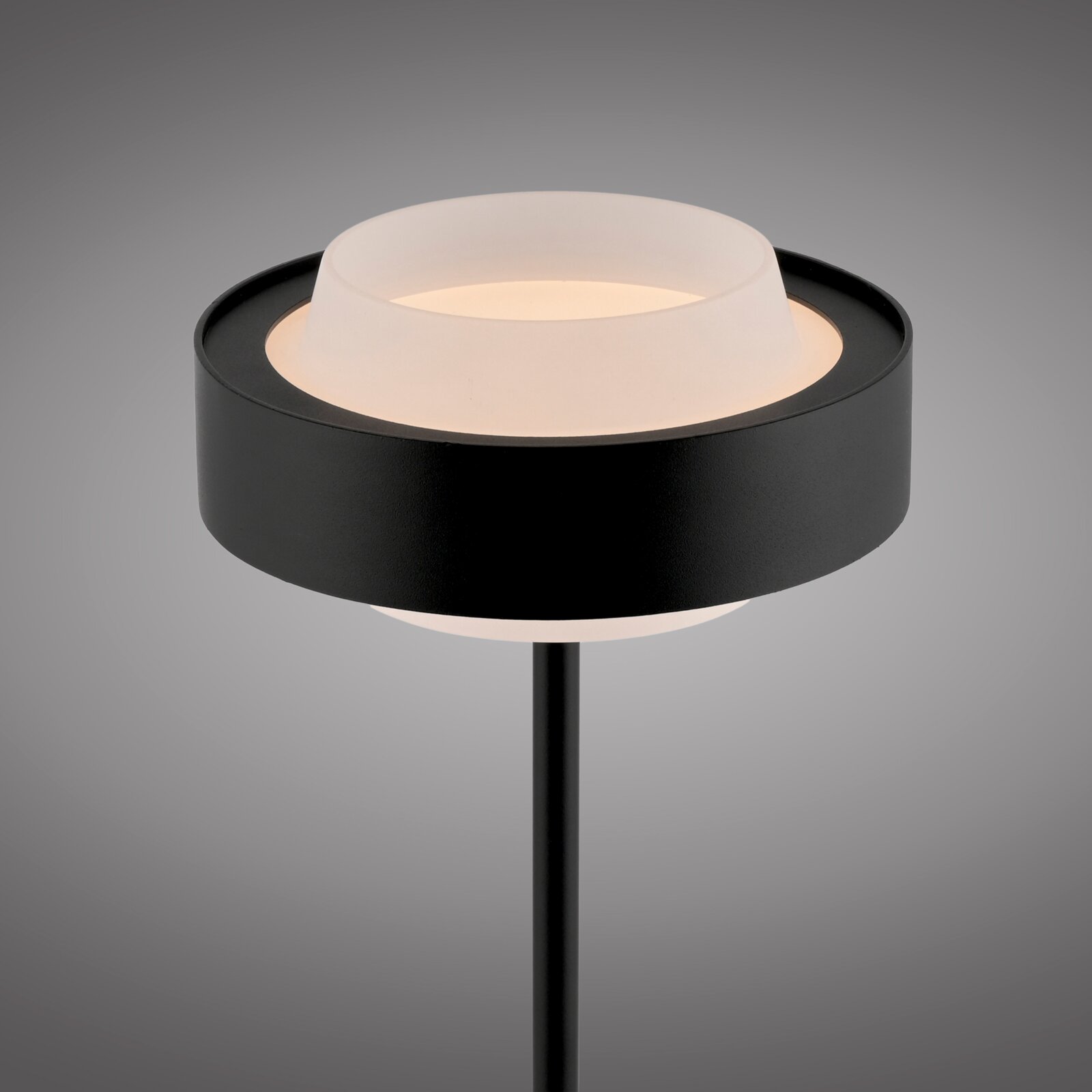 Moderne schwarze Außen-Stehleuchte mit ringförmigem Schirm und opalem Diffusor, warmweiß leuchtend, Nahaufnahme von schräg oben vor grauem Hintergrund