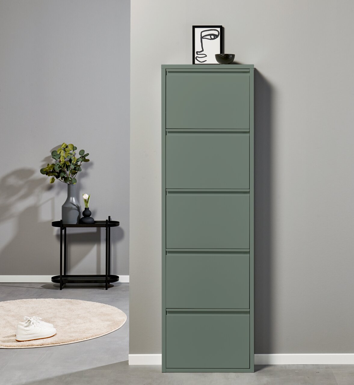 Schmaler, hoher Schuhschrank in mattem Salbeigrün mit fünf Klappfächern, frontal aus leicht erhöhter Perspektive vor heller Wand; oben Deko mit Bild und Schale.