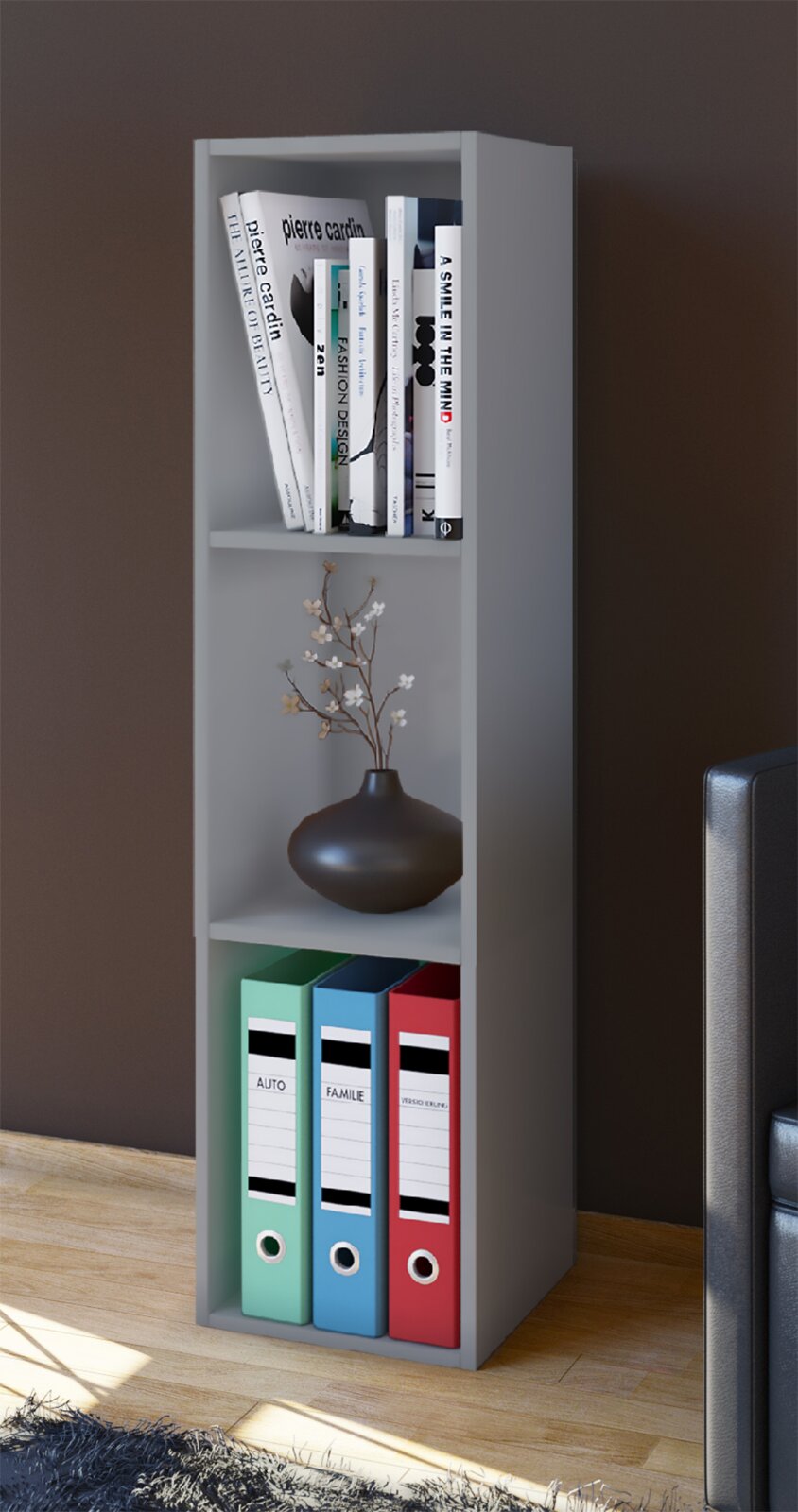 Schmales graues Standregal mit drei Fächern, oben Bücher, mittig eine dunkle Vase mit Zweigen, unten drei farbige Ordner; Ansicht leicht schräg von vorne.