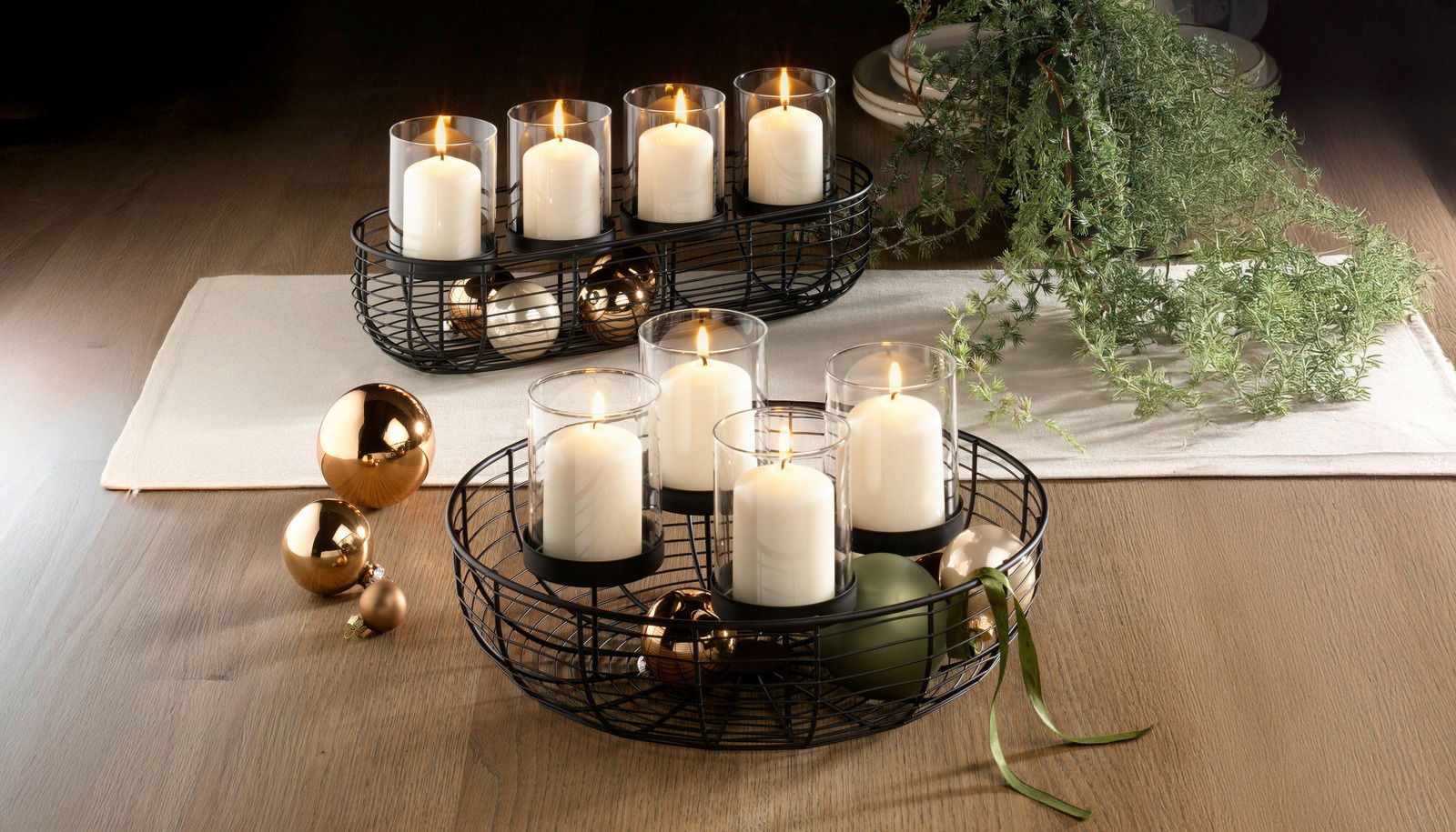 Dekorativer runder Kranz mit vier weißen Kerzen in Glaszylindern, umgeben von goldenen und grünen Kugeln, auf einem Holztisch von oben betrachtet.