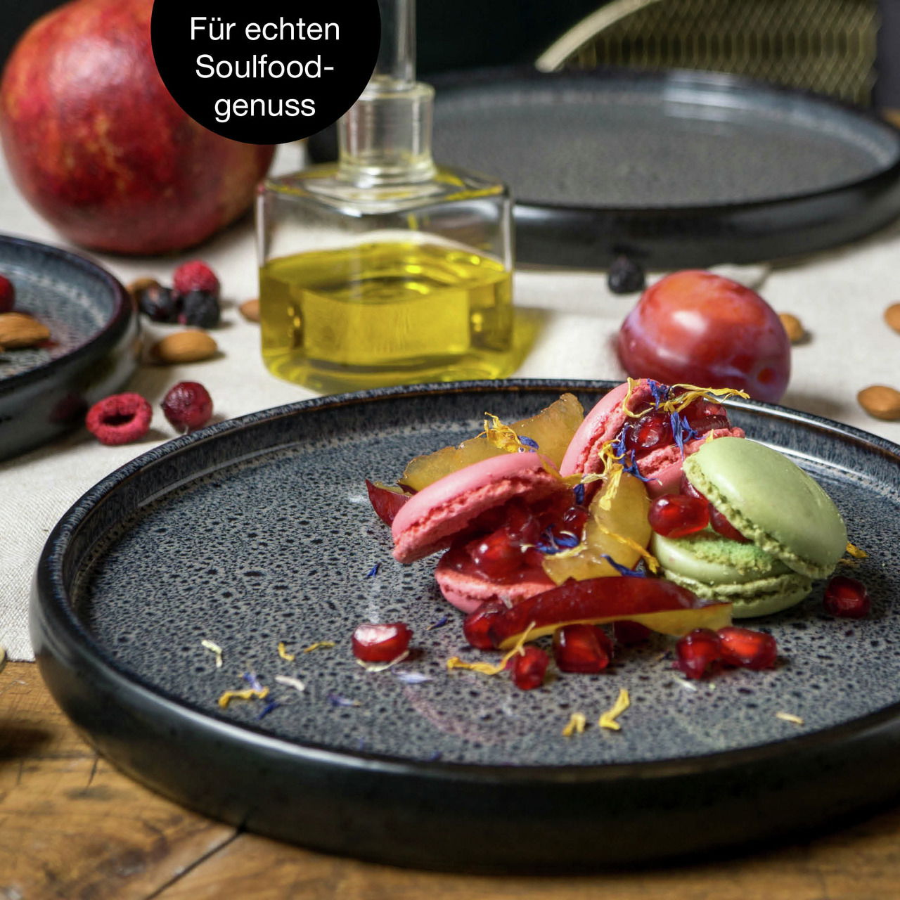 Nahaufnahme eines grauen Tellers mit Macarons und Früchten, umgeben von weiteren Tellern und Zutaten auf einem Holztisch.