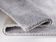 Nahaufnahme eines weichen, grauen Teppichs mit sichtbarer Rückseite, perspektivisch von der Seite aufgenommen.