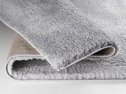 Nahaufnahme eines weichen, grauen Teppichs mit sichtbarer Rückseite, perspektivisch von der Seite aufgenommen.