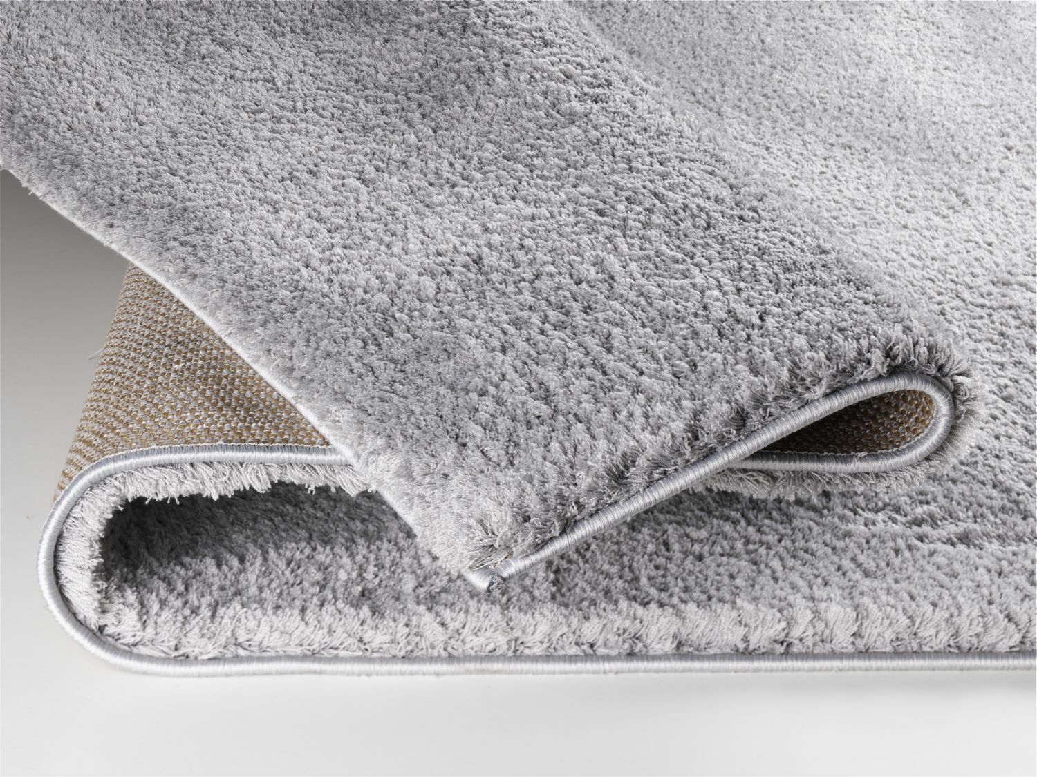 Nahaufnahme eines weichen, grauen Teppichs mit sichtbarer Rückseite, perspektivisch von der Seite aufgenommen.
