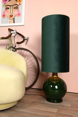 Grüne Stehlampe mit rundem Sockel und zylindrischem Schirm, seitliche Perspektive, neben einem gelben Sessel und einem Fahrrad vor einer rosa Wand.