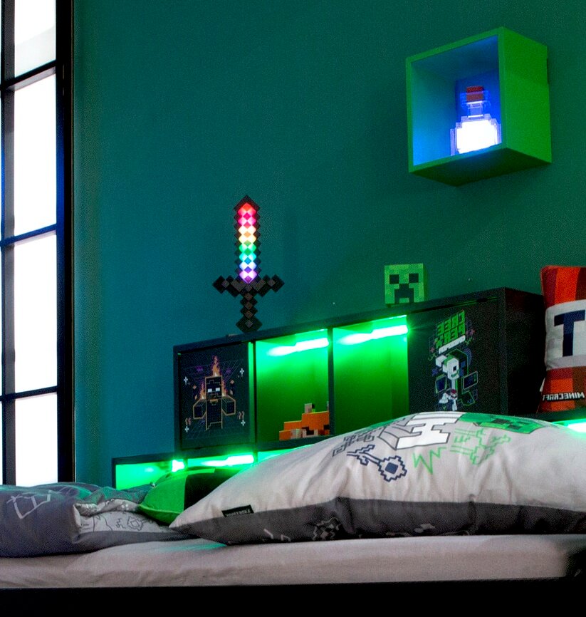 Minecraft-inspiriertes Kinderzimmer mit Bett und grün leuchtendem LED-Kopfteil, Pixel-Schwert und Deko an der Wand, aufgenommen aus seitlicher Perspektive auf Betthöhe.