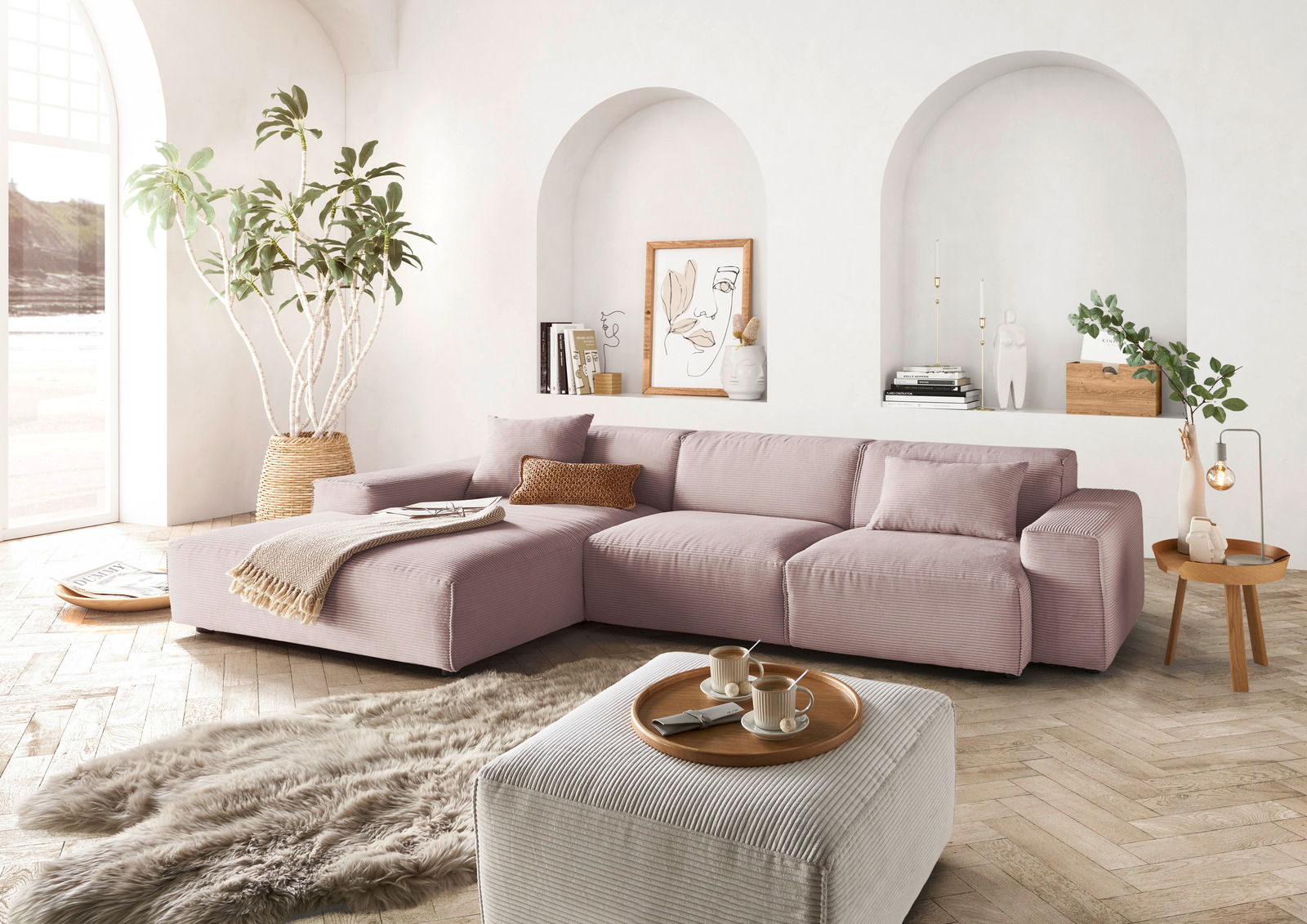 Gemütliche Wohnzimmer-Szene mit einer großen, modernen Eckcouch in sanftem Rosa, dekoriert mit Kissen und einer Decke. Die Couch steht in einem hellen Raum mit großen Fenstern, Pflanzen und stilvollen Dekorationen. Perspektive von vorne links.