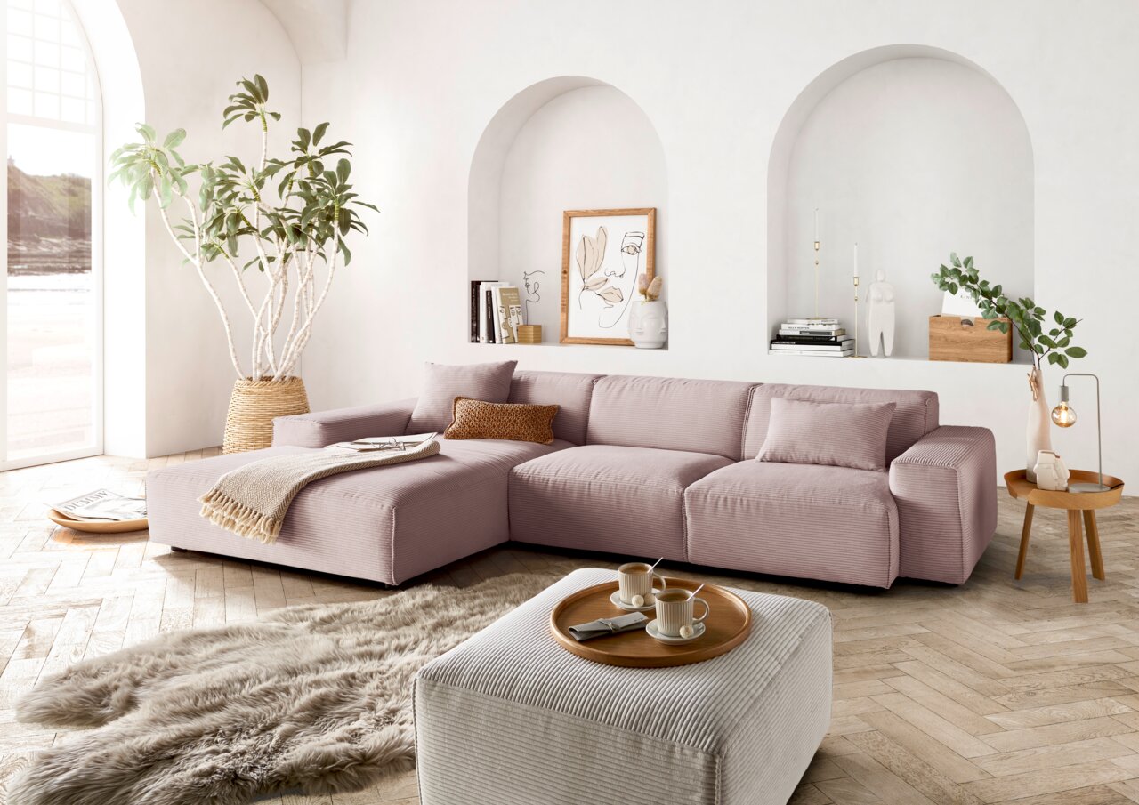 Gemütliche Wohnzimmer-Szene mit einer großen, modernen Eckcouch in sanftem Rosa, dekoriert mit Kissen und einer Decke. Die Couch steht in einem hellen Raum mit großen Fenstern, Pflanzen und stilvollen Dekorationen. Perspektive von vorne links.
