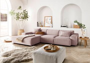 Gemütliche Wohnzimmer-Szene mit einer großen, modernen Eckcouch in sanftem Rosa, dekoriert mit Kissen und einer Decke. Die Couch steht in einem hellen Raum mit großen Fenstern, Pflanzen und stilvollen Dekorationen. Perspektive von vorne links.