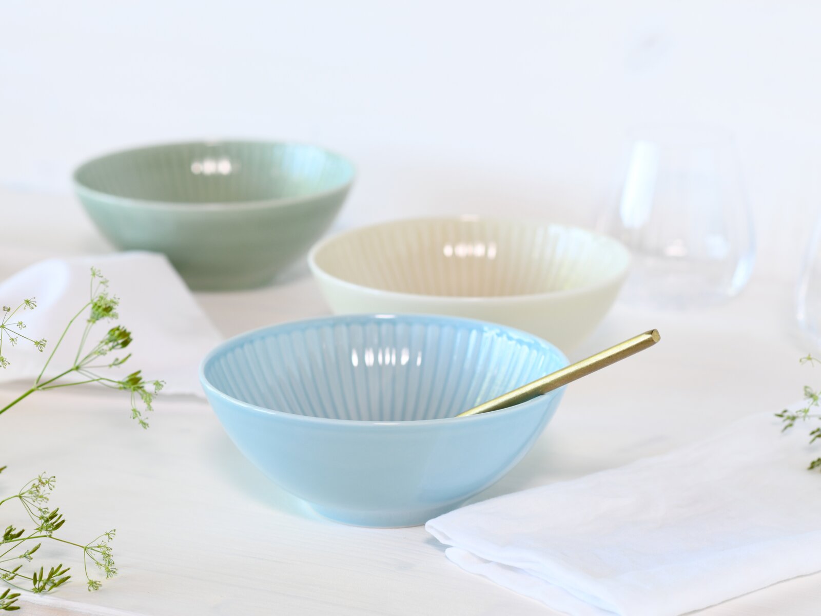 Pastellblaue Keramikschale mit geriffelter Innenstruktur und goldfarbenem Löffel auf weißem Tisch, im Hintergrund weitere Schalen in Grün und Creme; Perspektive: leicht seitliche Vorderansicht auf Tischhöhe
