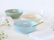 Pastellblaue Keramikschale mit geriffelter Innenstruktur und goldfarbenem Löffel auf weißem Tisch, im Hintergrund weitere Schalen in Grün und Creme; Perspektive: leicht seitliche Vorderansicht auf Tischhöhe