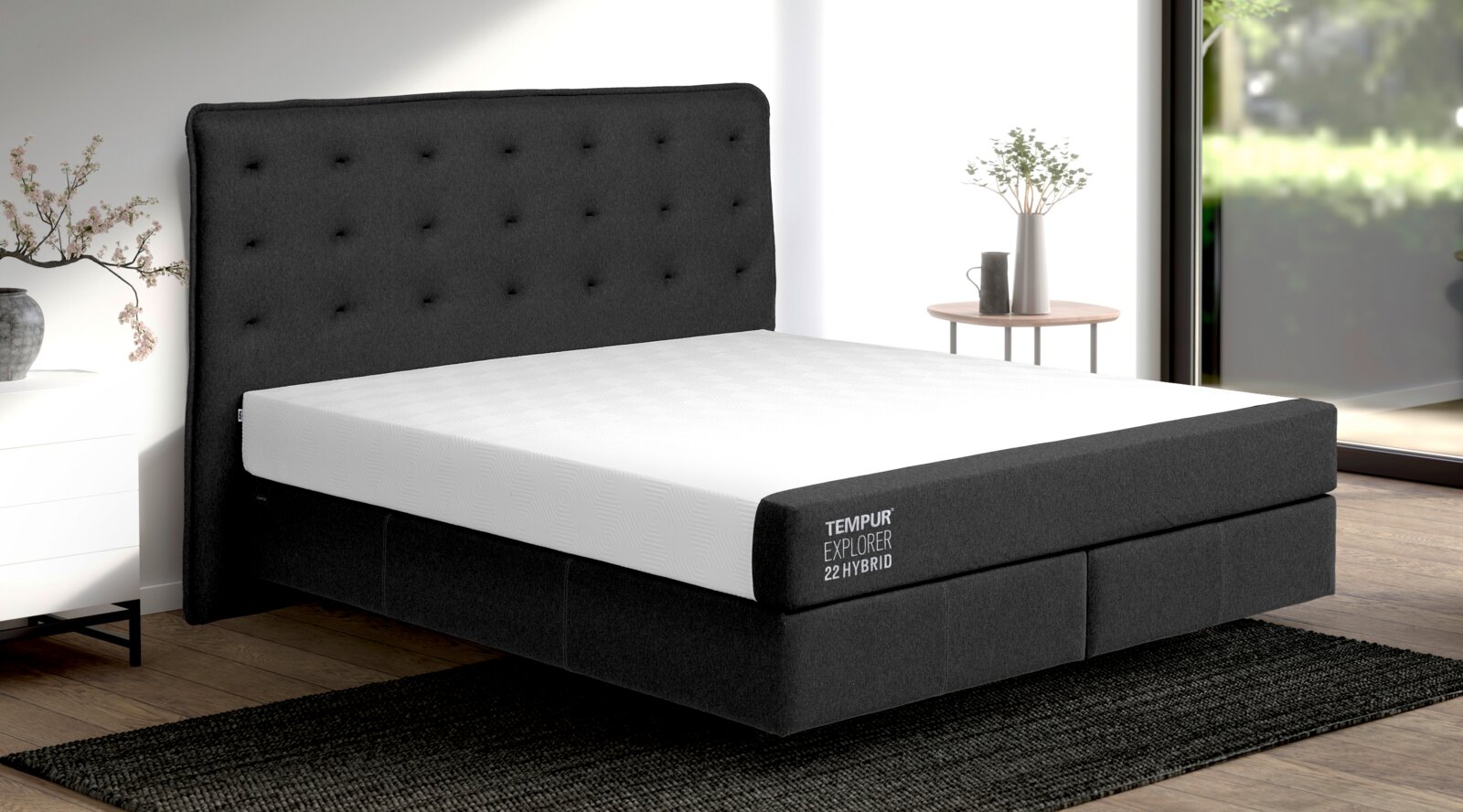 Boxspringbett mit grauem, gepolstertem Kopfteil und weißer Matratze, seitliche Perspektive