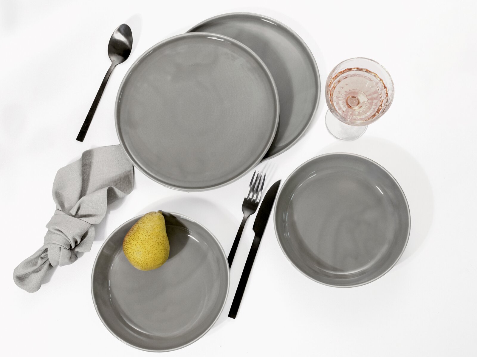 Graues Keramikgeschirr-Set mit mehreren Tellern, Schale, schwarzem Besteck, Stoffserviette und Weinglas auf weißem Hintergrund, Draufsicht von oben
