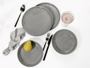 Graues Keramikgeschirr-Set mit mehreren Tellern, Schale, schwarzem Besteck, Stoffserviette und Weinglas auf weißem Hintergrund, Draufsicht von oben