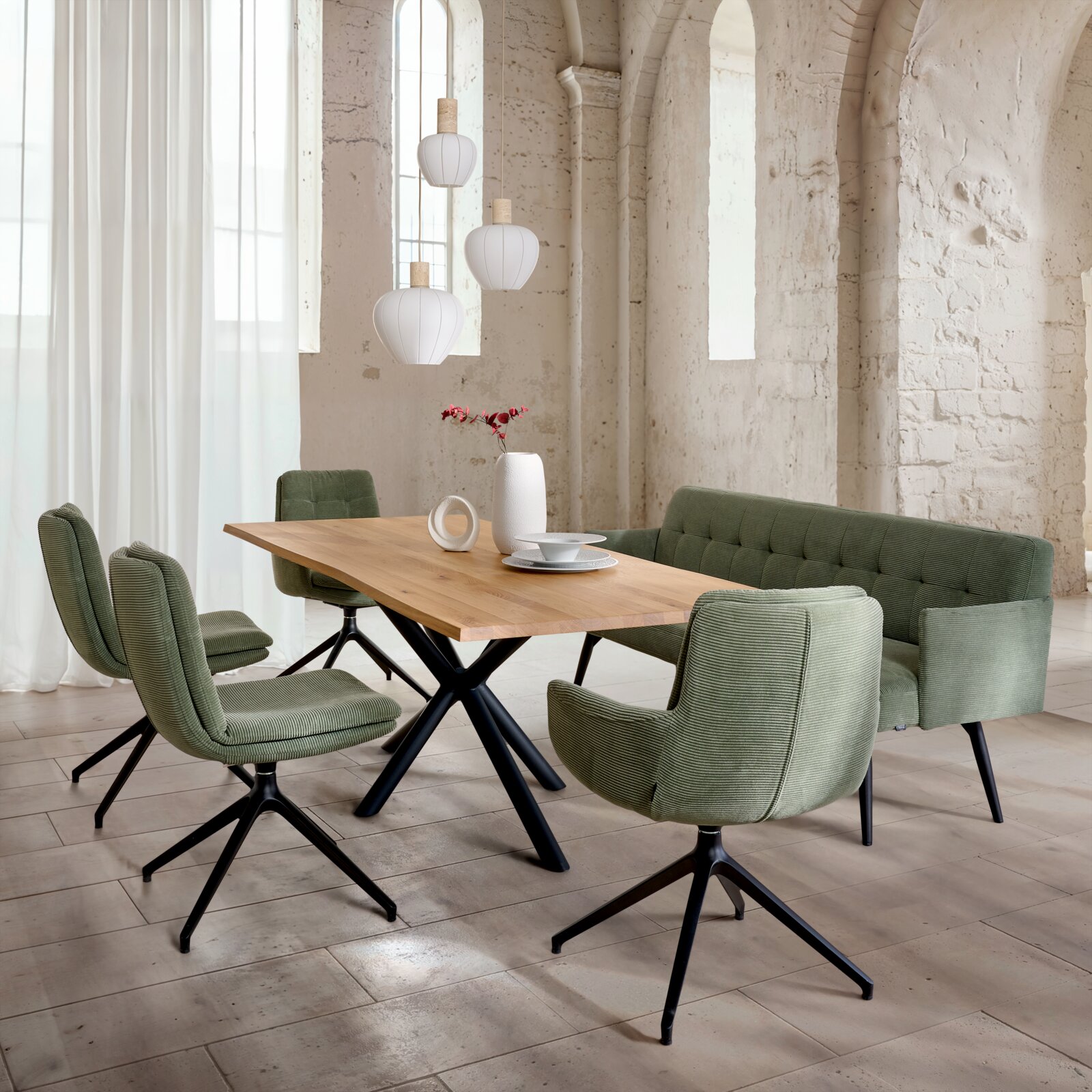 Schräg von vorn fotografierte Essgruppe: rechteckiger Holztisch mit schwarzem Metallgestell, grüne Polsterbank und zwei grüne Drehstühle in hellem Loft mit hängenden weißen Lampen.