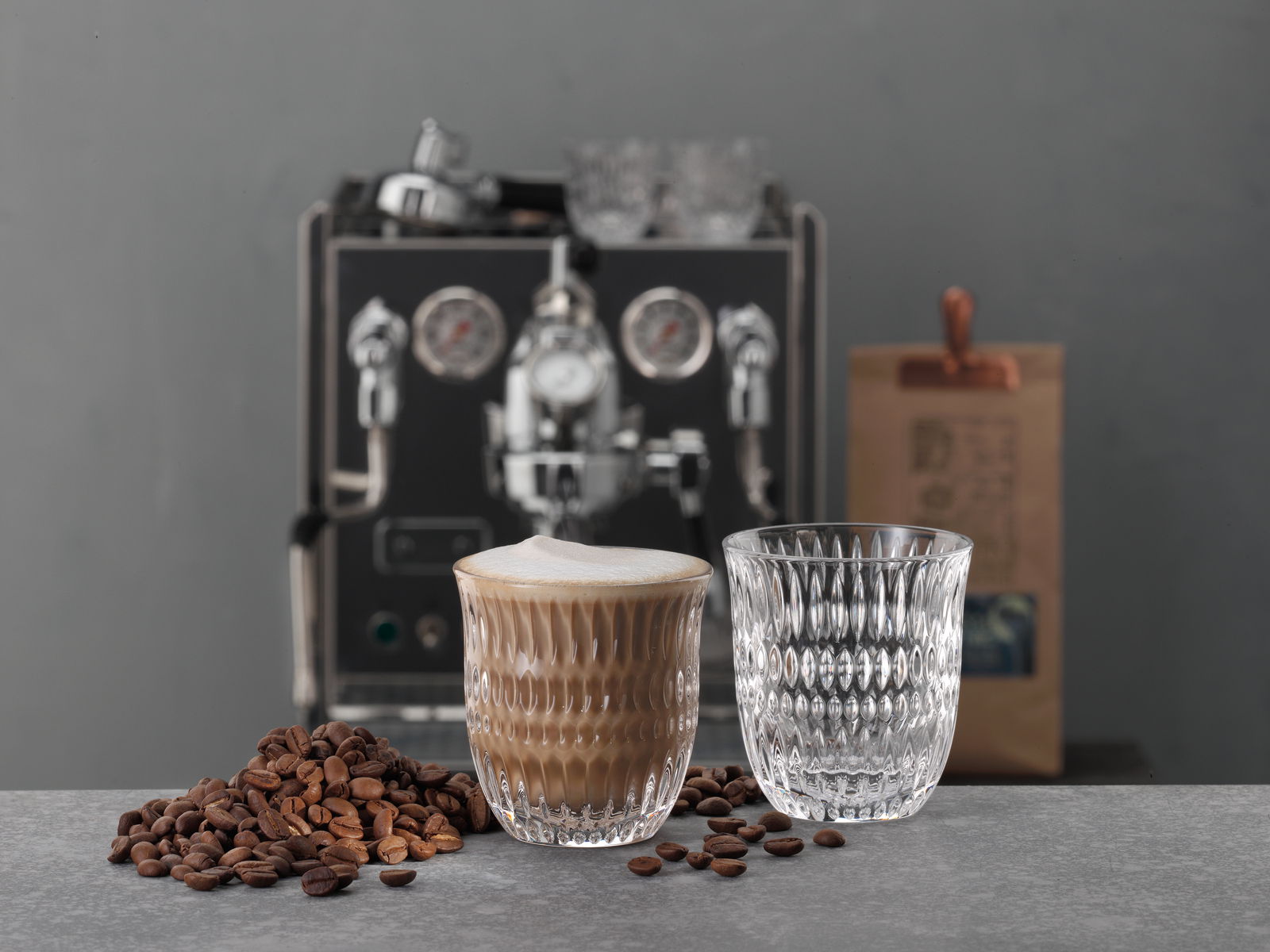Zwei elegante Gläser, eines mit Cappuccino gefüllt, auf einem Tisch mit Kaffeebohnen, im Hintergrund eine Kaffeemaschine und eine Kaffeeverpackung. Perspektive: seitlich.