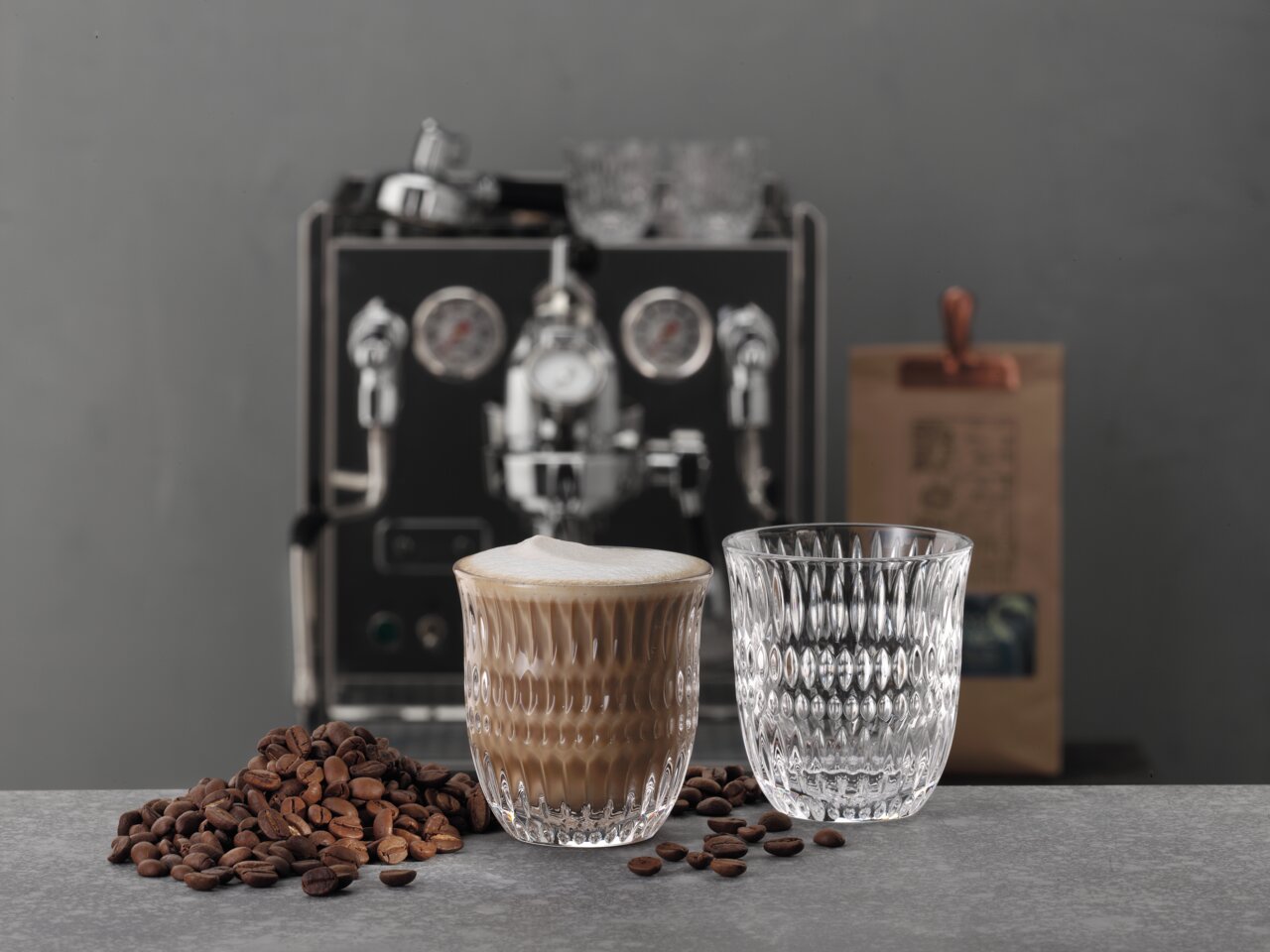 Twee elegante glazen, waarvan één gevuld met cappuccino, op een tafel met koffiebonen, op de achtergrond een koffiezetapparaat en een verpakking koffie. Perspectief: zijaanzicht.