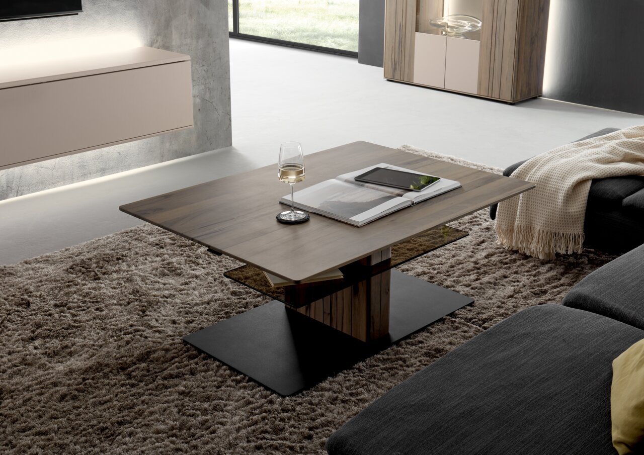 Moderner Couchtisch aus Holz mit Glasablage, seitliche Perspektive, dekoriert mit einem Weinglas und einem Buch auf einem Teppichboden.
