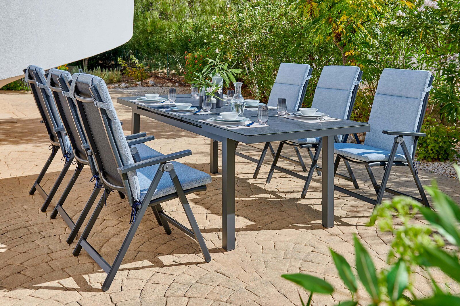 Sechs blaue Klappsessel um einen gedeckten Gartentisch auf einer Terrasse, seitliche Perspektive