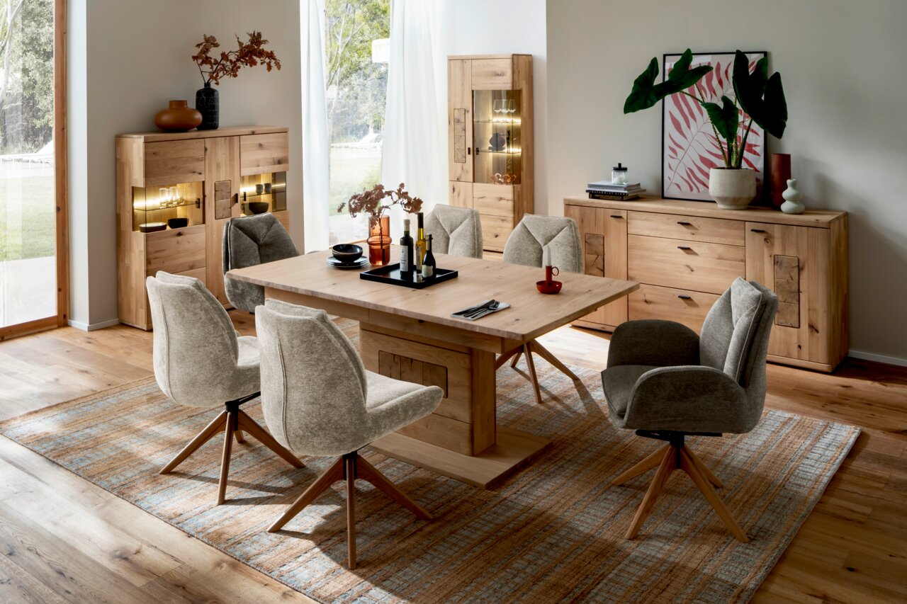 Esszimmer mit Holztisch und sechs gepolsterten Stühlen in einem modernen, hellen Raum. Die Perspektive zeigt den Tisch von der Seite, umgeben von Stühlen auf einem Teppich, mit Schränken und Dekorationen im Hintergrund.
