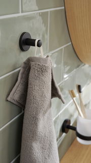 Schwarzer Handtuchhaken von tesa MOON BLACK an einer gefliesten Wand, seitliche Perspektive, mit einem grauen Handtuch aufgehängt.