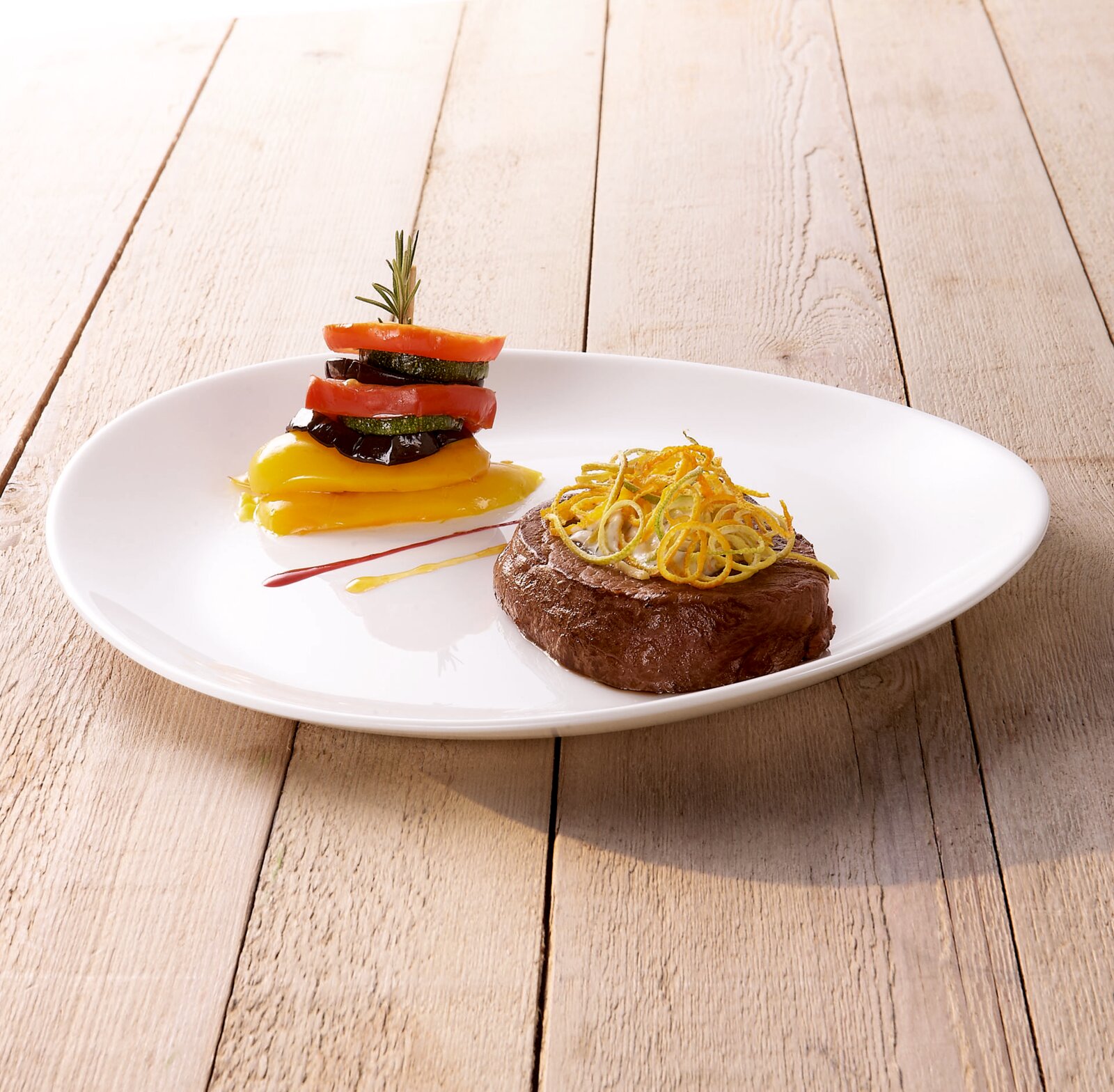 Gourmet-Teller mit gebratenem Steak und knusprigen Gemüsestreifen sowie gestapeltem Ratatouille-Gemüse auf gelbem Püree, auf weißem ovalen Teller auf Holztisch, aufgenommen aus leicht schräger Seitenansicht (3/4-Perspektive)