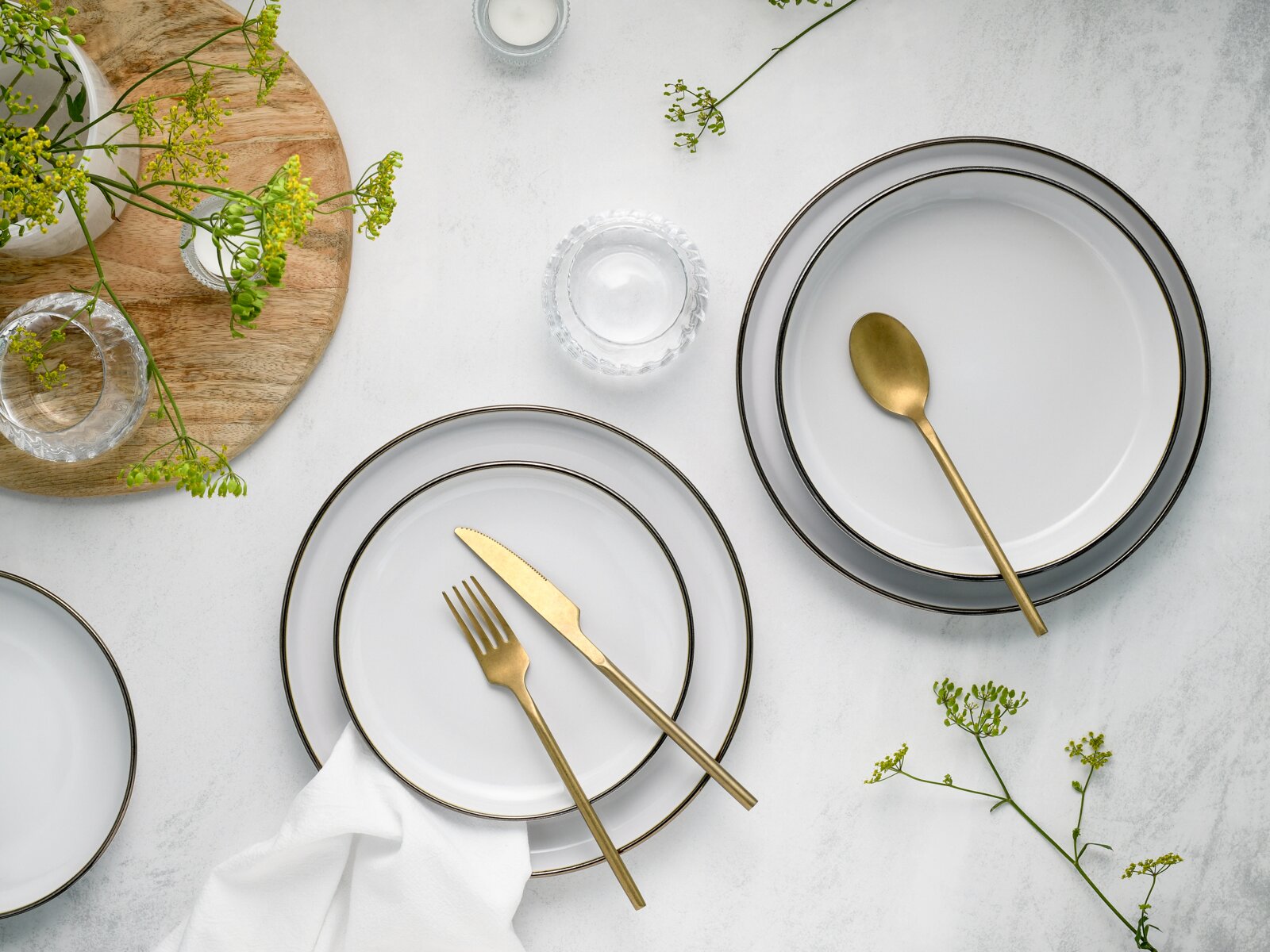 Draufsicht auf ein elegantes Tischgedeck mit weißen Tellern mit dunklem Rand, goldfarbenem Besteck, Glas-Schälchen und grünen Blütenzweigen auf heller Steinoberfläche