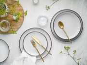 Draufsicht auf ein elegantes Tischgedeck mit weißen Tellern mit dunklem Rand, goldfarbenem Besteck, Glas-Schälchen und grünen Blütenzweigen auf heller Steinoberfläche