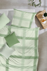 Bettwäsche-Set in zartem Grün mit dezentem Muster und passenden Kissen auf weißem Bett, aufgenommen aus der Vogelperspektive (Top-Down) neben einem Holz-Nachttisch mit Pflanze und Gebäck.