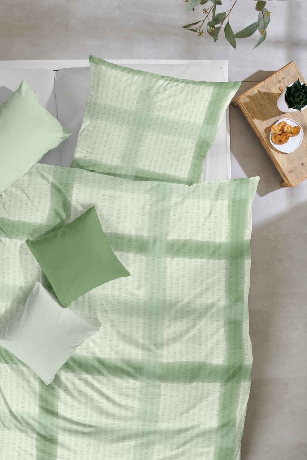 Bettwäsche-Set in zartem Grün mit dezentem Muster und passenden Kissen auf weißem Bett, aufgenommen aus der Vogelperspektive (Top-Down) neben einem Holz-Nachttisch mit Pflanze und Gebäck.