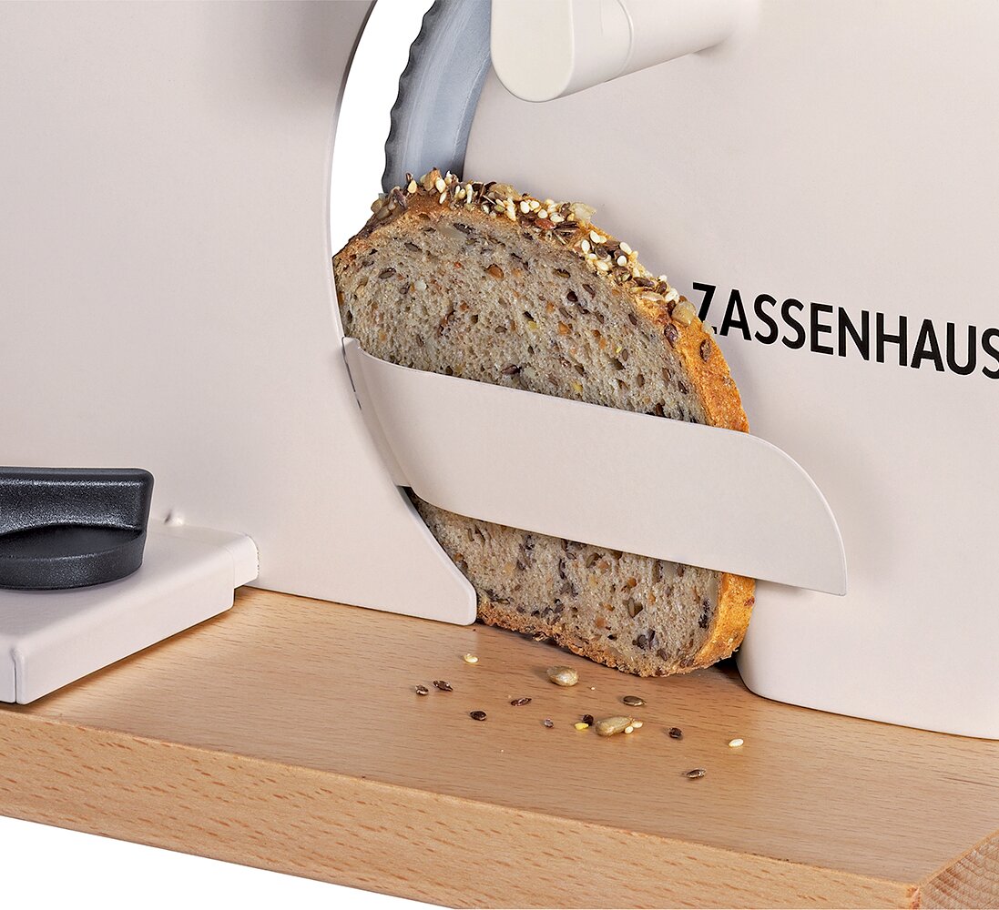 Close-up zijaanzicht van een witte Zassenhaus broodsnijmachine die een plak meergranenbrood snijdt op een houten snijplank, met enkele zaden en kruimels ervoor
