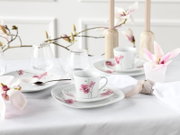 Elegant gedeckter Tisch mit weißem Porzellan-Kaffeegeschirr mit rosa Magnolienblüten, Glasbechern und Kerzenhaltern, fotografiert aus leicht erhöhter schräger Vorderansicht.