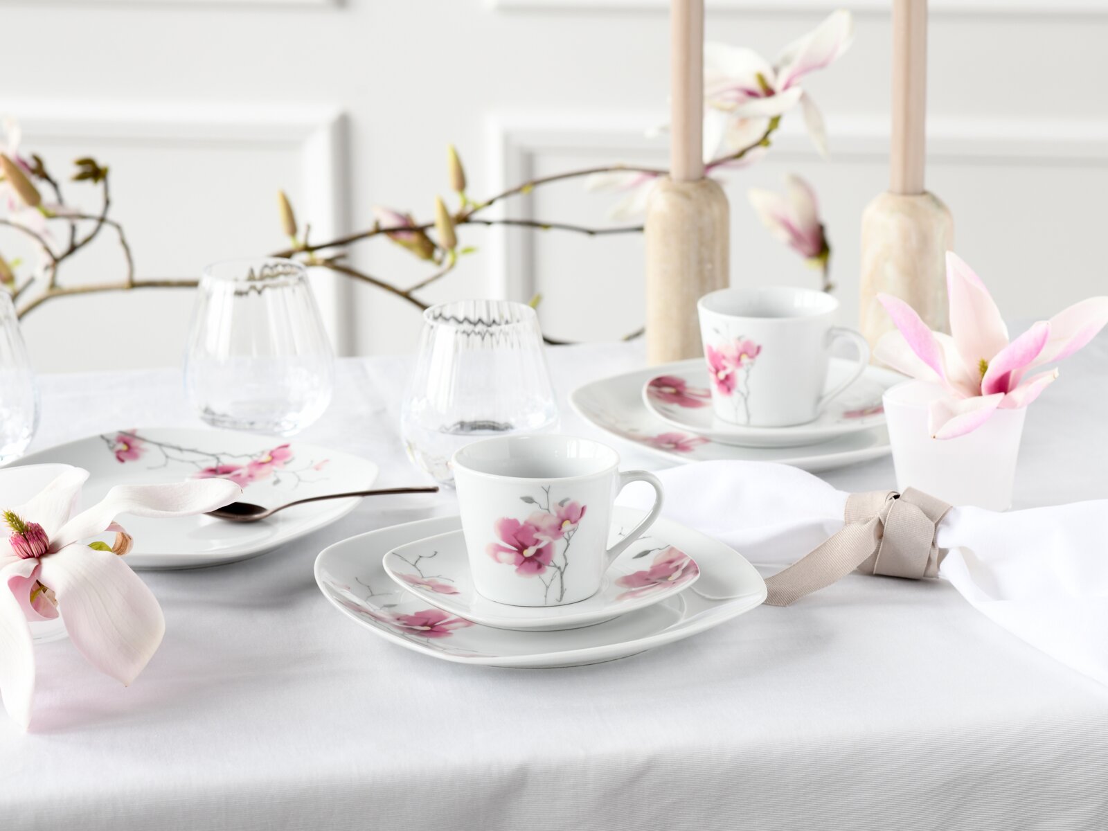 Elegant gedeckter Tisch mit weißem Porzellan-Kaffeegeschirr mit rosa Magnolienblüten, Glasbechern und Kerzenhaltern, fotografiert aus leicht erhöhter schräger Vorderansicht.