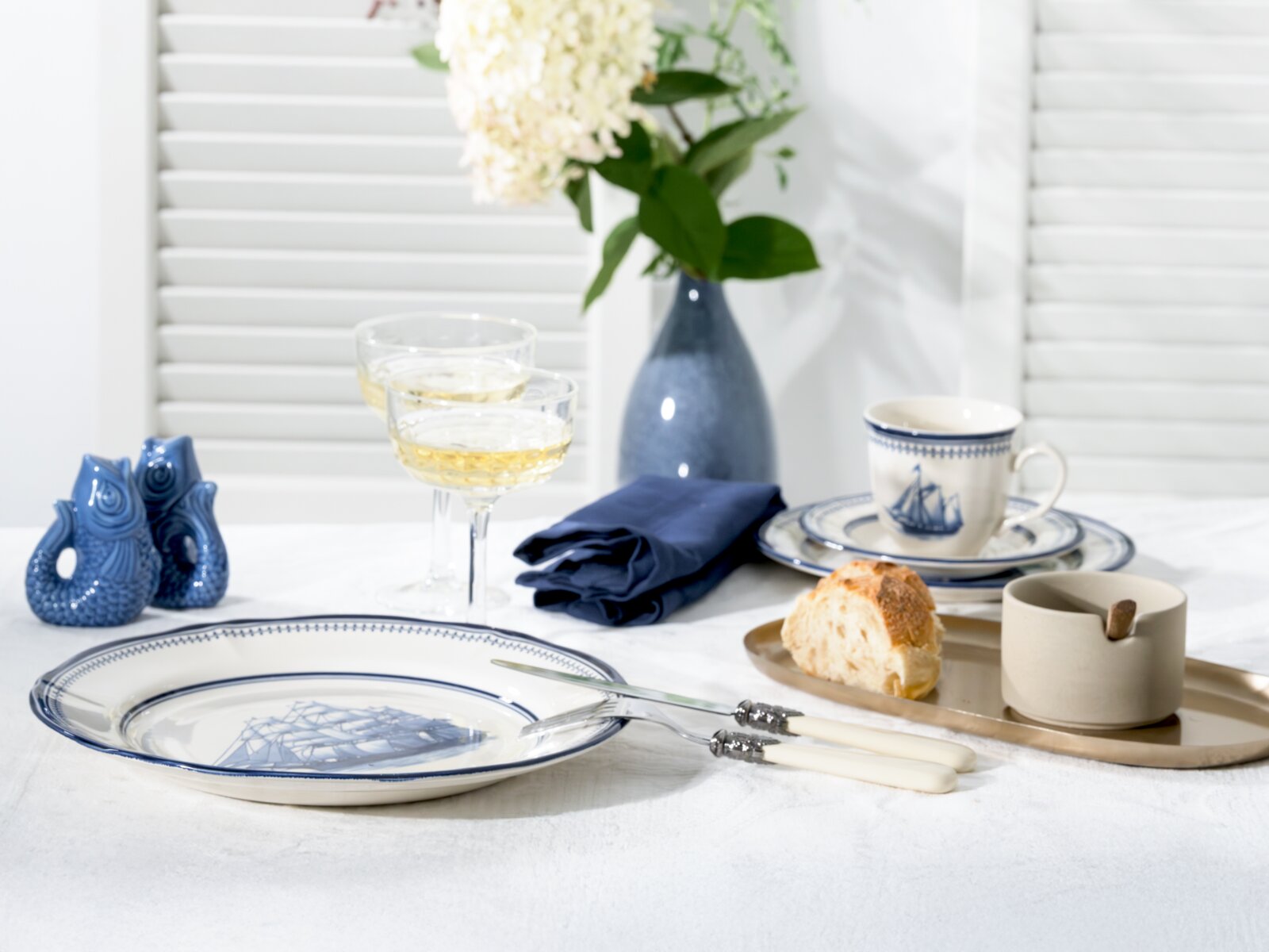 Maritim gedeckter Tisch mit blau-weißem Porzellanservice (Teller mit Schiffsmotiv, Tasse und Untertasse), zwei Champagnergläsern, blauem Stoffserviette, Messer mit hellen Griffen, Brot auf Tablett sowie blauer Vase mit weißen Blumen; Perspektive: leicht v
