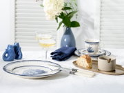 Maritim gedeckter Tisch mit blau-weißem Porzellanservice (Teller mit Schiffsmotiv, Tasse und Untertasse), zwei Champagnergläsern, blauem Stoffserviette, Messer mit hellen Griffen, Brot auf Tablett sowie blauer Vase mit weißen Blumen; Perspektive: leicht v
