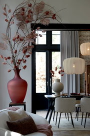 Ein stilvolles Wohnzimmer mit einem großen roten Blumenstrauß in einer Vase, einem Esstisch mit Stühlen und einer modernen Hängelampe. Im Vordergrund liegt ein beiges Kissen auf einem Sofa. Perspektive: seitlich von vorne.