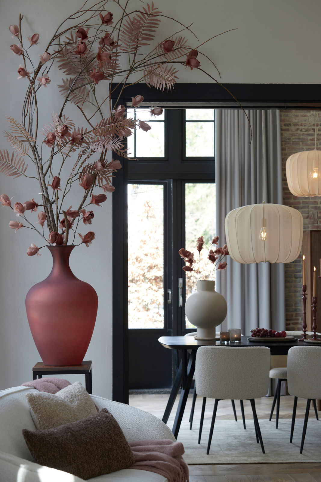 Ein stilvolles Wohnzimmer mit einem großen roten Blumenstrauß in einer Vase, einem Esstisch mit Stühlen und einer modernen Hängelampe. Im Vordergrund liegt ein beiges Kissen auf einem Sofa. Perspektive: seitlich von vorne.