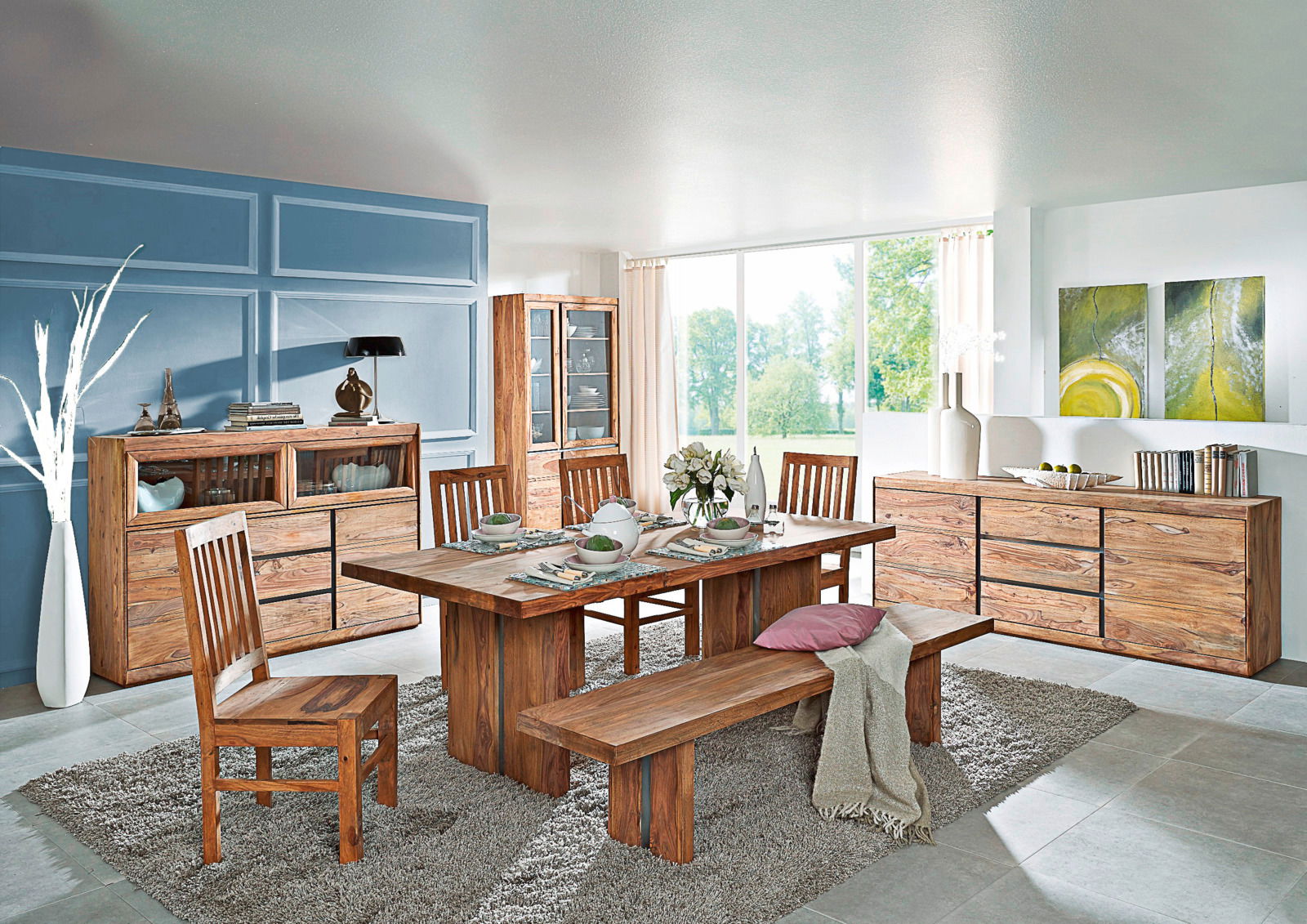 Esszimmer mit Holzmöbeln, bestehend aus einem großen Esstisch, Stühlen und einer Bank, umgeben von Sideboards und Vitrinen, mit Blick auf einen Garten durch große Fenster.